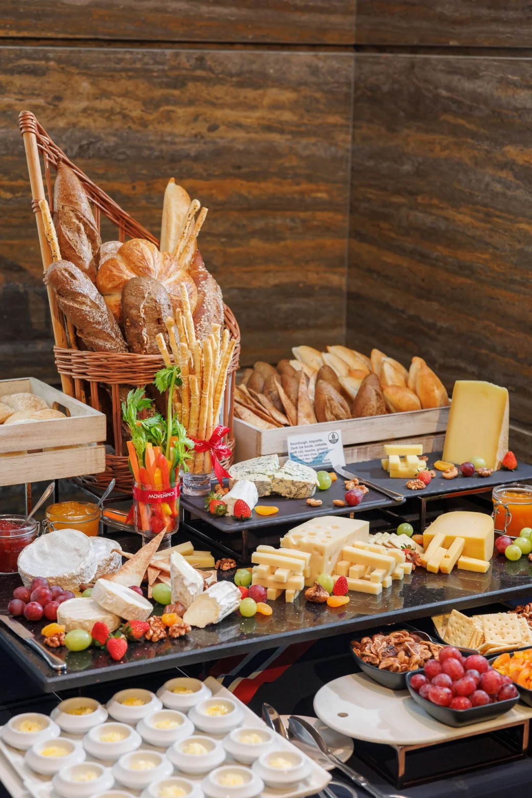 Barra de buffet servida: canasta de panes, tabla de quesos, uvas, fresas, nueces y frutos secos.